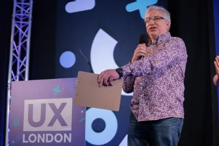 A man on stage with a microphone and a clipboard in front of a podium that reads: UX London.