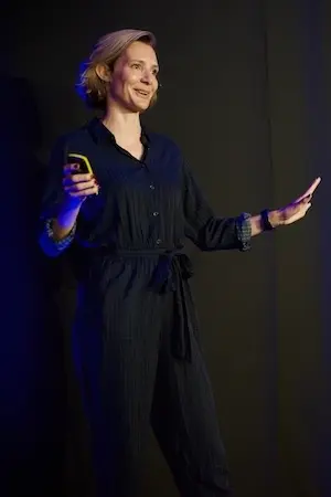 A woman speaking on stage, smiling and holding her arms out.