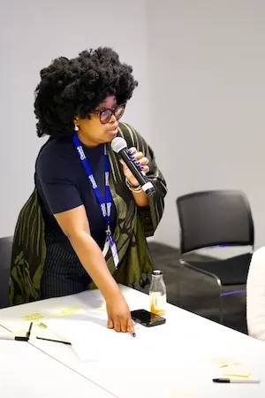 A woman in a workshop speaking with a microphone in one hand while she points to what's on the table.
