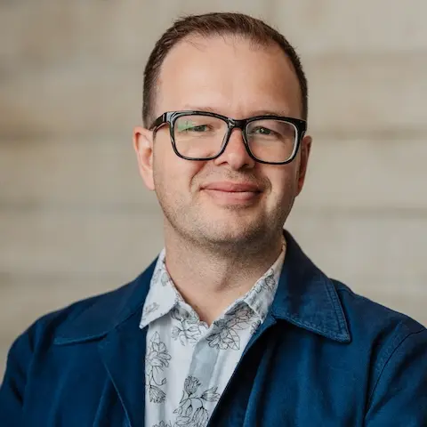 A smiling man with short hair and glasses wearing a smart-looking shirt and jacket.