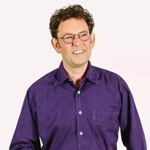 A smiling man with curly dark hair and glasses wearing a purple shirt.