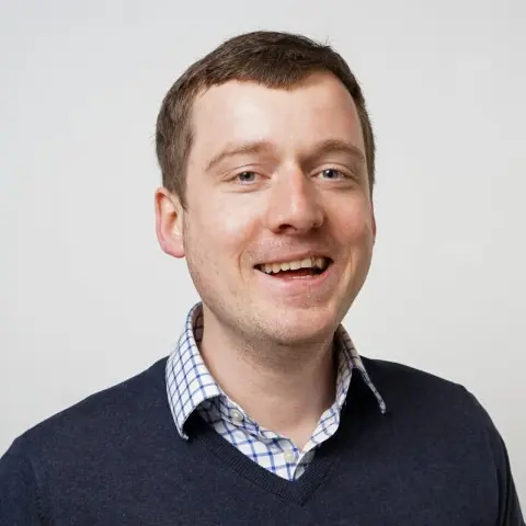 A smiling man with short hair wearing a collared shirt under his jumper