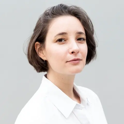 A woman facing to the side but with her head turned to the camera, wearing a white shirt against a grey background.