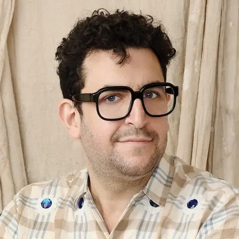 A man with short curly hair and glasses wearing a light plaid shirt in front of a light background