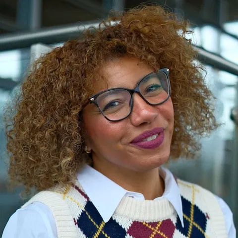 A woman with curly hair and glasses smiling and tilting her head.