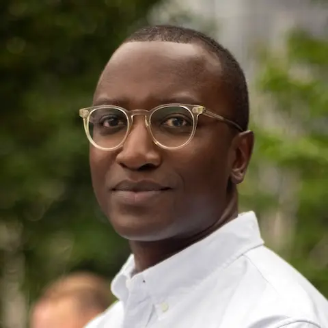 A man with short hair and glasses wearing a white shirt turning towards the camera.