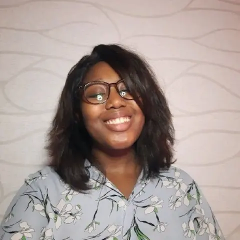 A smiling woman with glasses and shoulder-length hair wearing a floral top in front of a patterned background.