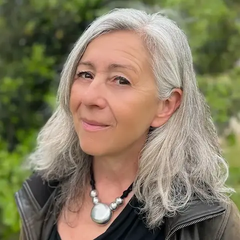 A woman with shoulder-length white hair and a jacket outdoors standing to the side and looking at the camera.